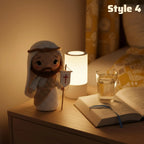 A crocheted Jesus figurine wearing a white robe and brown beard, holding a small cross banner, styled on a bedside table next to a glowing lamp, an open book, and a glass of water in a warm bedroom scene.