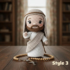 A crocheted doll of Jesus with a brown beard, white robe, and beige head covering, standing on a wooden table with a rosary in front. The background shows a softly blurred indoor room with bookshelves.