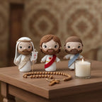 Three crocheted Jesus figurines in white robes with brown hair and beards, arranged on a wooden table with a rosary and a lit candle. The background is a softly blurred warm-toned interior.