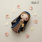 A crocheted doll styled as the Virgin Mary, wearing a navy blue robe with gold trim and a brown dress, lying on a light beige textured background with scattered pink and white flower petals. The text 'Style 5' is visible in the top right corner.