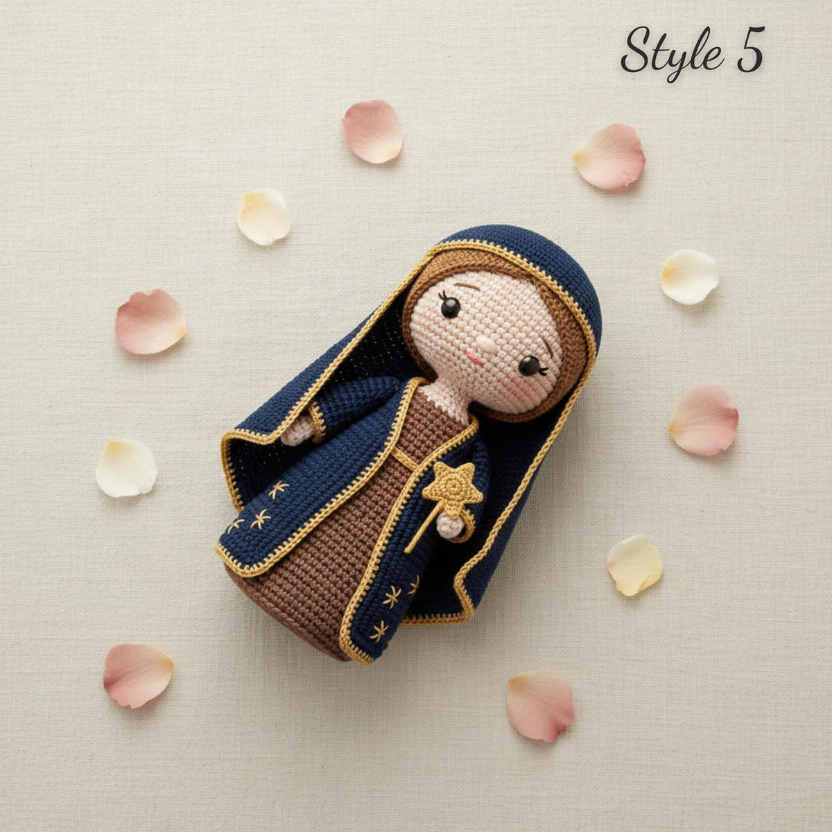 A crocheted doll styled as the Virgin Mary, wearing a navy blue robe with gold trim and a brown dress, lying on a light beige textured background with scattered pink and white flower petals. The text 'Style 5' is visible in the top right corner.