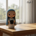 A crocheted doll of the Virgin Mary stands on a light wood table in front of a bright window with sheer curtains. The doll has a blue and cream hood, a cream dress, and holds a black book with a gold cross.