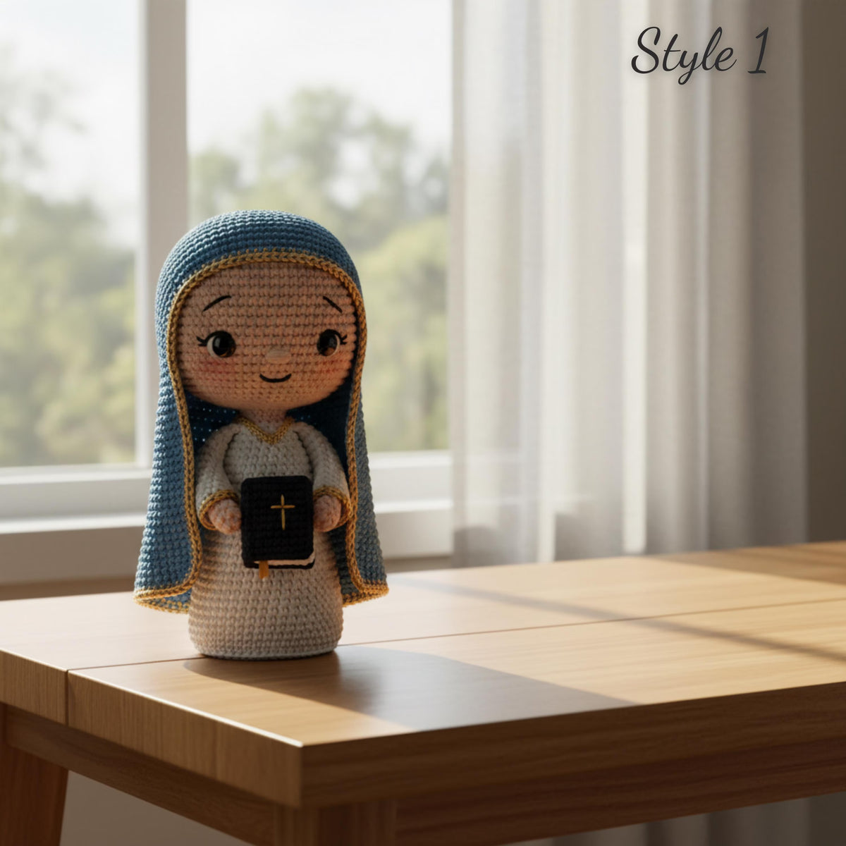 A crocheted doll of the Virgin Mary stands on a light wood table in front of a bright window with sheer curtains. The doll has a blue and cream hood, a cream dress, and holds a black book with a gold cross.