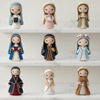 Nine crocheted doll figurines arranged on white shelves against a light background. The dolls are dressed in various colored robes, including blue, pink, green, navy, gold, and brown, and each has a large embroidered face.