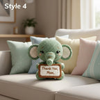 A crocheted elephant plush toy sits on a light beige sofa surrounded by pastel pillows. The plush is mint green with cream inner ears, embroidered facial features, and holds a small sign reading 'Thank You Mom.