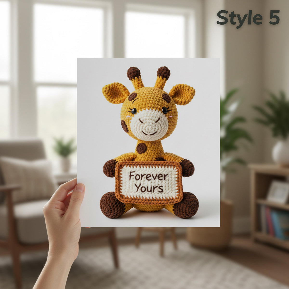 A hand holds a printed card or poster featuring a crocheted giraffe holding a sign that says 'Forever Yours,' with the text 'Style 5' in the top right. The background shows a softly blurred living room interior with a chair, windows, and plants.