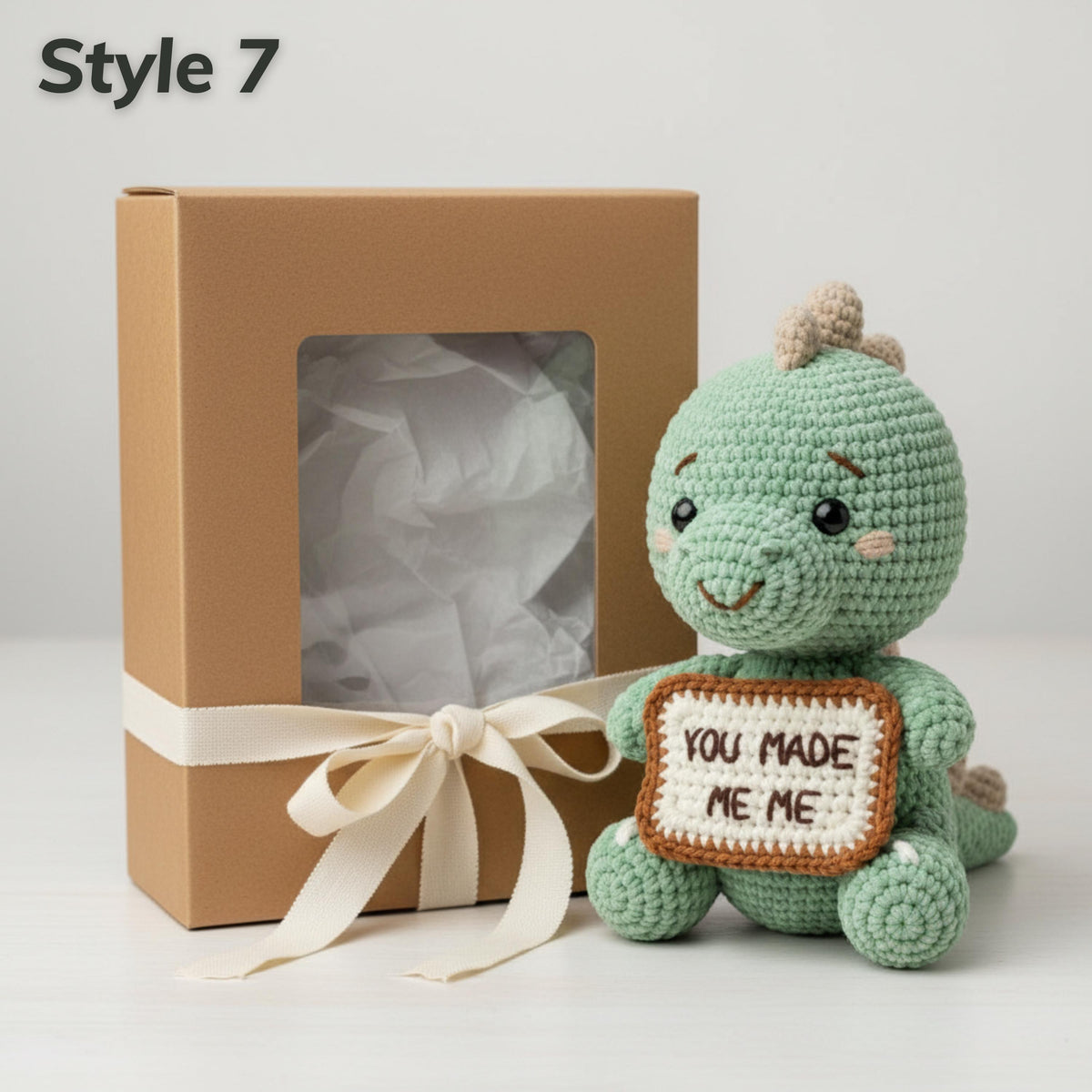 A crocheted green dinosaur plush toy holding a sign that says 'YOU MADE ME ME' sits next to a brown gift box with a cream ribbon, on a light gray background. The text 'Style 7' is in the top-left corner.