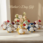Six crocheted penguin toys in different colors are arranged on a light wooden surface with a soft beige background and a blurred floral arrangement behind them. The text 'Mother's Day Gift' appears at the top.