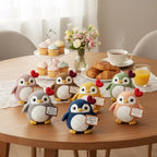 A group of crocheted penguin figurines in multiple colors (pink, brown, yellow, green, blue, peach) arranged on a wooden table, each holding a small sign that reads 'Thank You for Eating With Us'. The scene is styled as a cozy cafe or bakery table with cupcakes, pastries, a coffee cup, and an orange glass in the background.