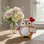 A crocheted penguin toy sits on a wooden table in a softly lit room, with a vase of pink and white flowers behind it. The penguin is mauve pink with a white belly, yellow beak and feet, and a red heart on its head, holding a small sign that reads, 'Thank You For Everything, Mom.