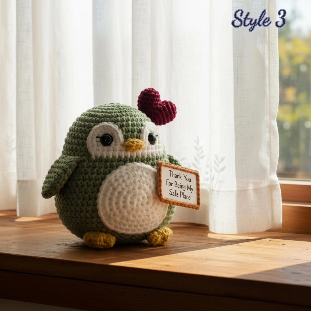 A crocheted penguin plush toy sits on a wooden windowsill with sheer white curtains and a window in the background. The penguin is olive green with a white belly, yellow beak and feet, and a dark red heart on its head.