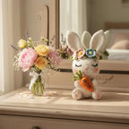 A crocheted white bunny doll wearing a flower crown and holding an orange carrot, placed on a light wood dresser beside a small glass vase with pink and yellow flowers.