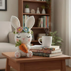 A crocheted white bunny plush holding an orange carrot sits on a wooden table in a cozy living room setting, next to a stack of books and a white mug with steam.