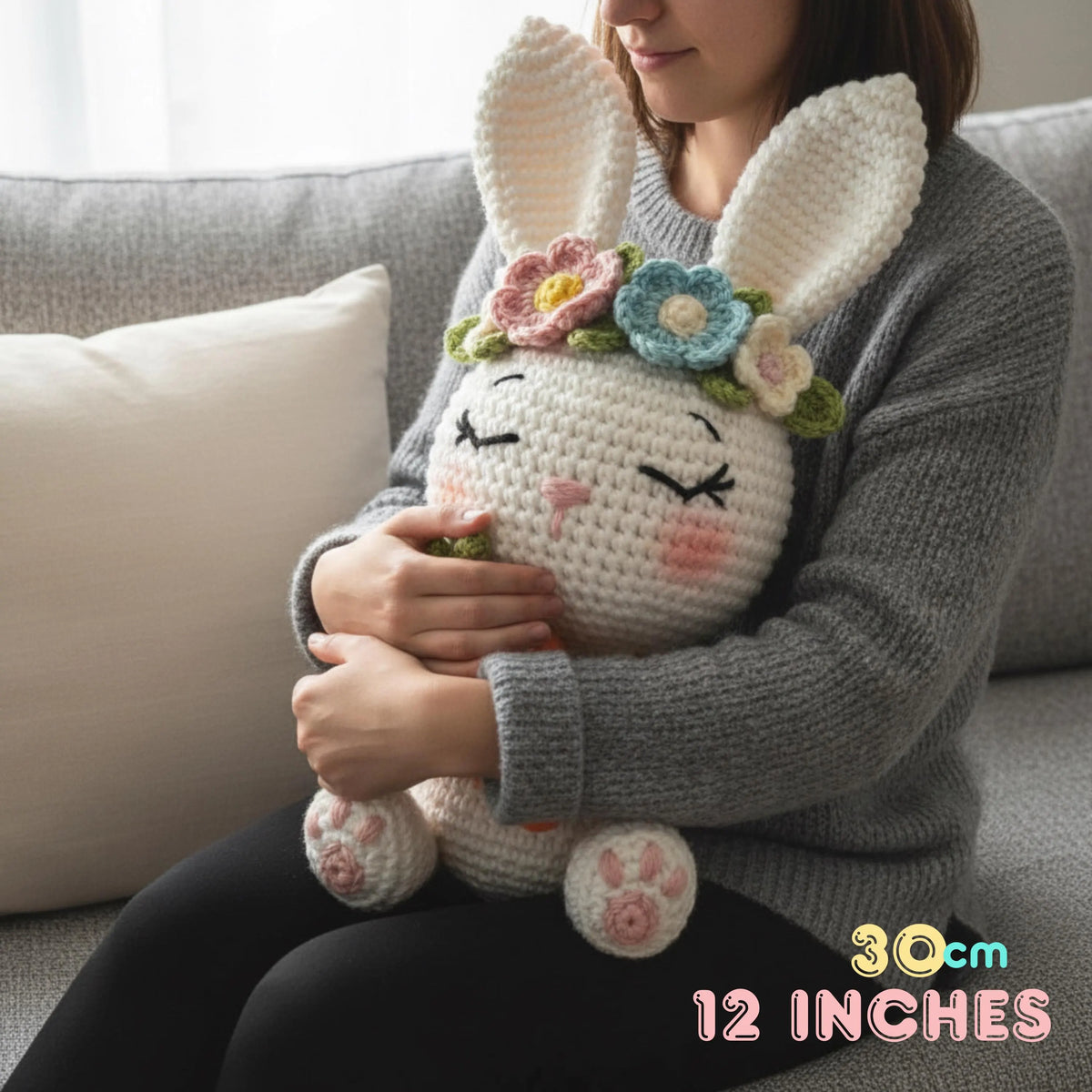 A woman in a gray knit sweater holds a large crocheted bunny plush toy with long white ears, closed embroidered eyes, pink cheeks, and pastel flowers on its head. The woman is seated on a light gray sofa with a cream pillow, and a text overlay in the bottom right reads '30cm 12 INCHES'.