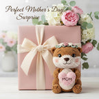 A pink gift box tied with a cream ribbon sits on a light surface, styled with a crocheted brown bear toy holding a pink heart that says “MOM,” surrounded by pink and white flowers. The words “Perfect Mother’s Day Surprise” appear at the top.