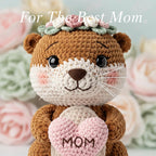 A crocheted otter plush toy with a floral crown and a pink heart that says 'MOM', centered against a soft blurred floral background with the text 'For The Best Mom' across the top.