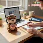 A crocheted otter plush toy sits on a wooden desk beside an open laptop, with a person writing in a notebook nearby. The otter has brown and cream fur, a pink heart with 'MOM' embroidered on the front, and a small flower crown.
