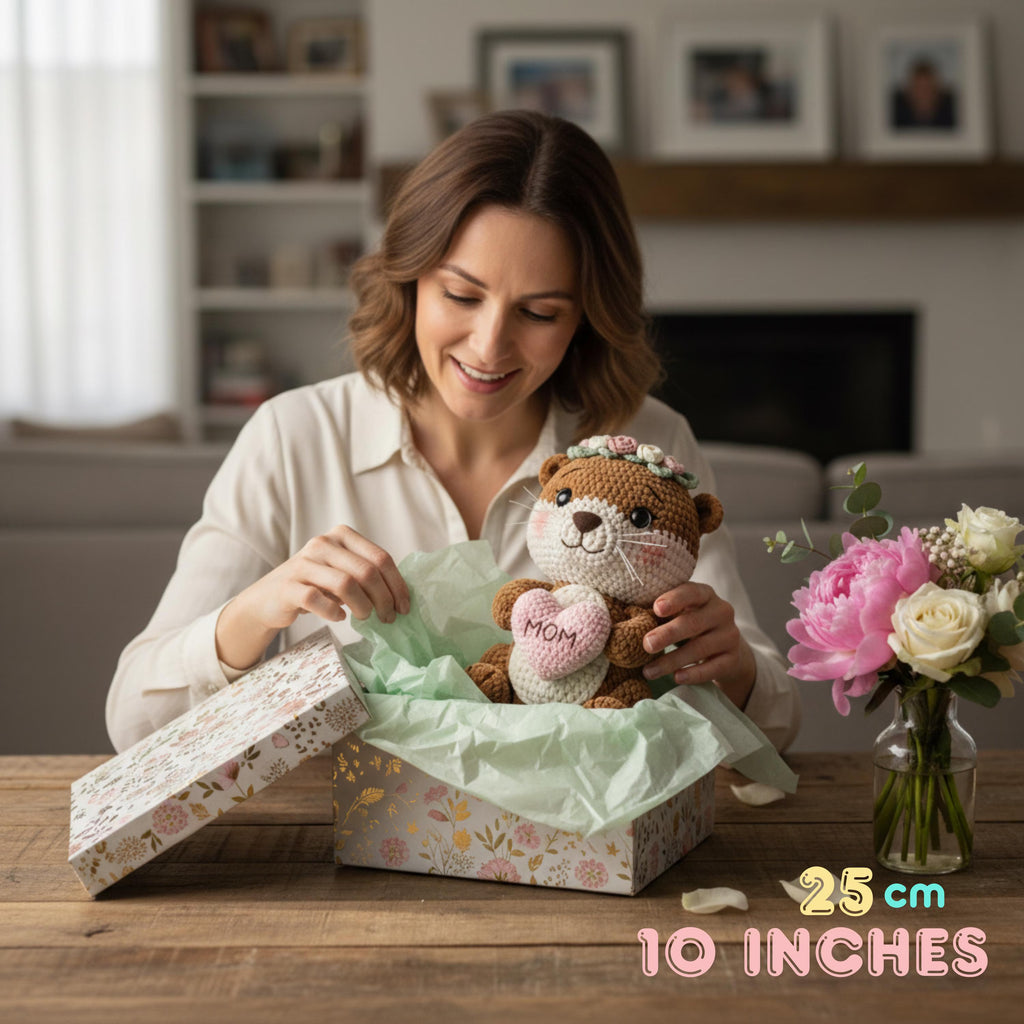 A woman in a cream shirt smiles while opening a floral gift box to reveal a crocheted teddy bear with a pink heart that says 'MOM'. The scene is set in a cozy living room with a vase of flowers on the table, and text in the corner reads '25 cm 10 INCHES'.