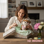 A woman in a cream shirt smiles while opening a floral gift box to reveal a crocheted teddy bear with a pink heart that says 'MOM'. The scene is set in a cozy living room with a vase of flowers on the table, and text in the corner reads '25 cm 10 INCHES'.