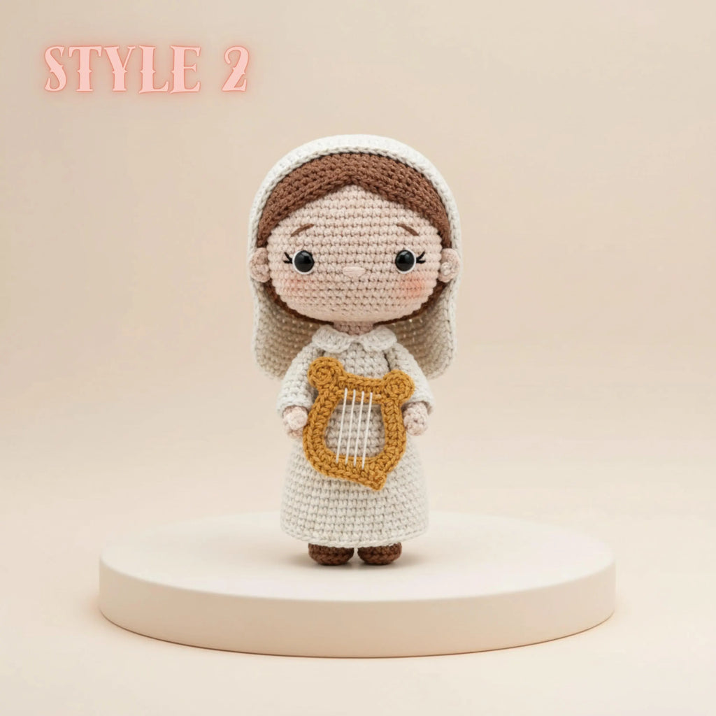 A crocheted doll figure with a white dress, brown hair, and a gold harp accessory, standing on a round pedestal against a warm beige background. The text 'STYLE 2' appears in the upper left corner.