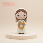 A crocheted doll figure with a white dress, brown hair, and a gold harp accessory, standing on a round pedestal against a warm beige background. The text 'STYLE 2' appears in the upper left corner.
