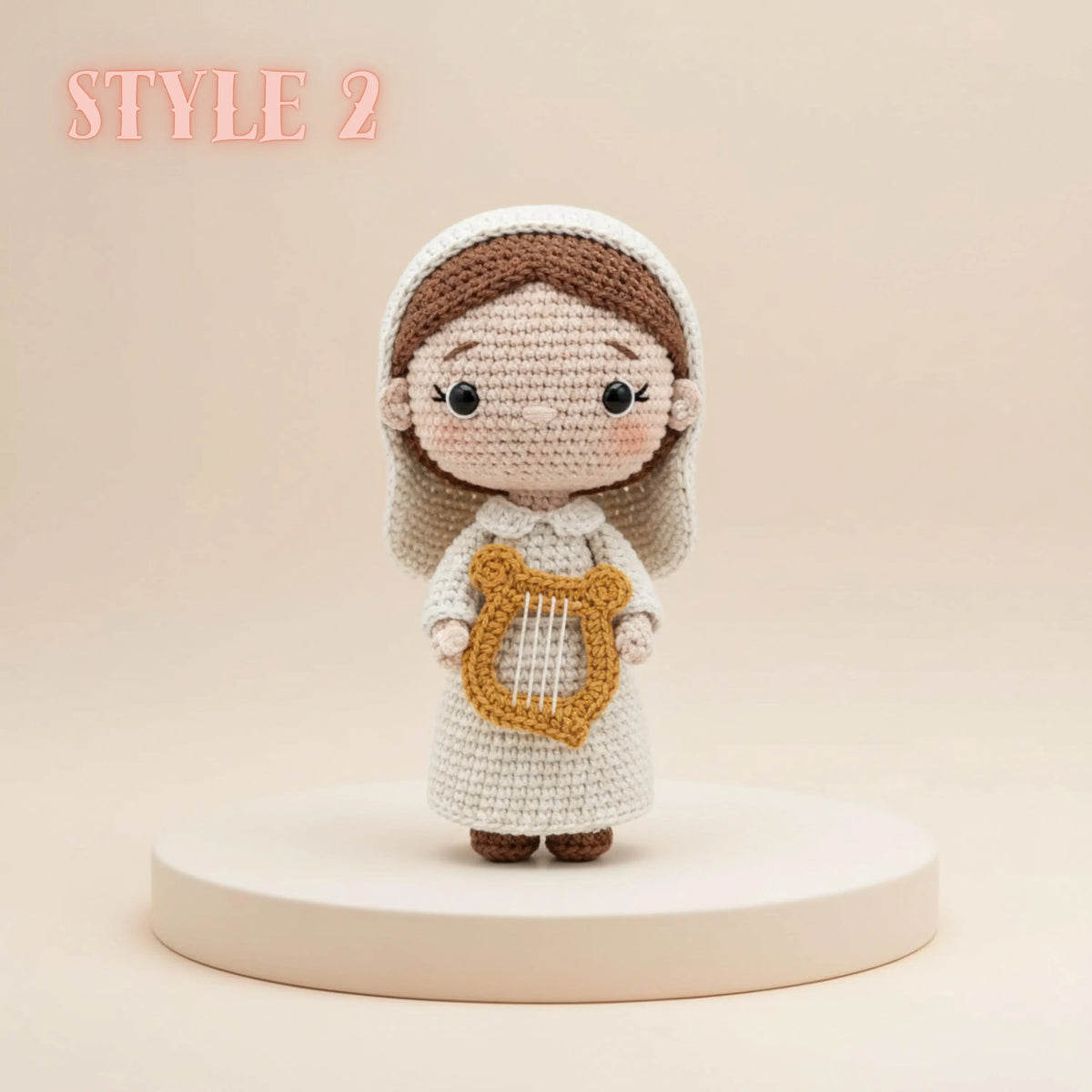 A crocheted doll figure with a white dress, brown hair, and a gold harp accessory, standing on a round pedestal against a warm beige background. The text 'STYLE 2' appears in the upper left corner.