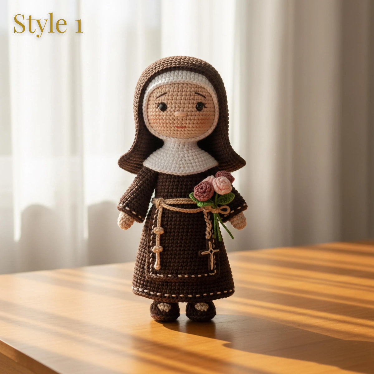 A crocheted doll dressed as a nun stands on a wooden table in front of soft white curtains. The doll wears a dark brown habit and dress with a white veil, has a rope belt with a cross, and holds a small bouquet of pink and mauve roses.