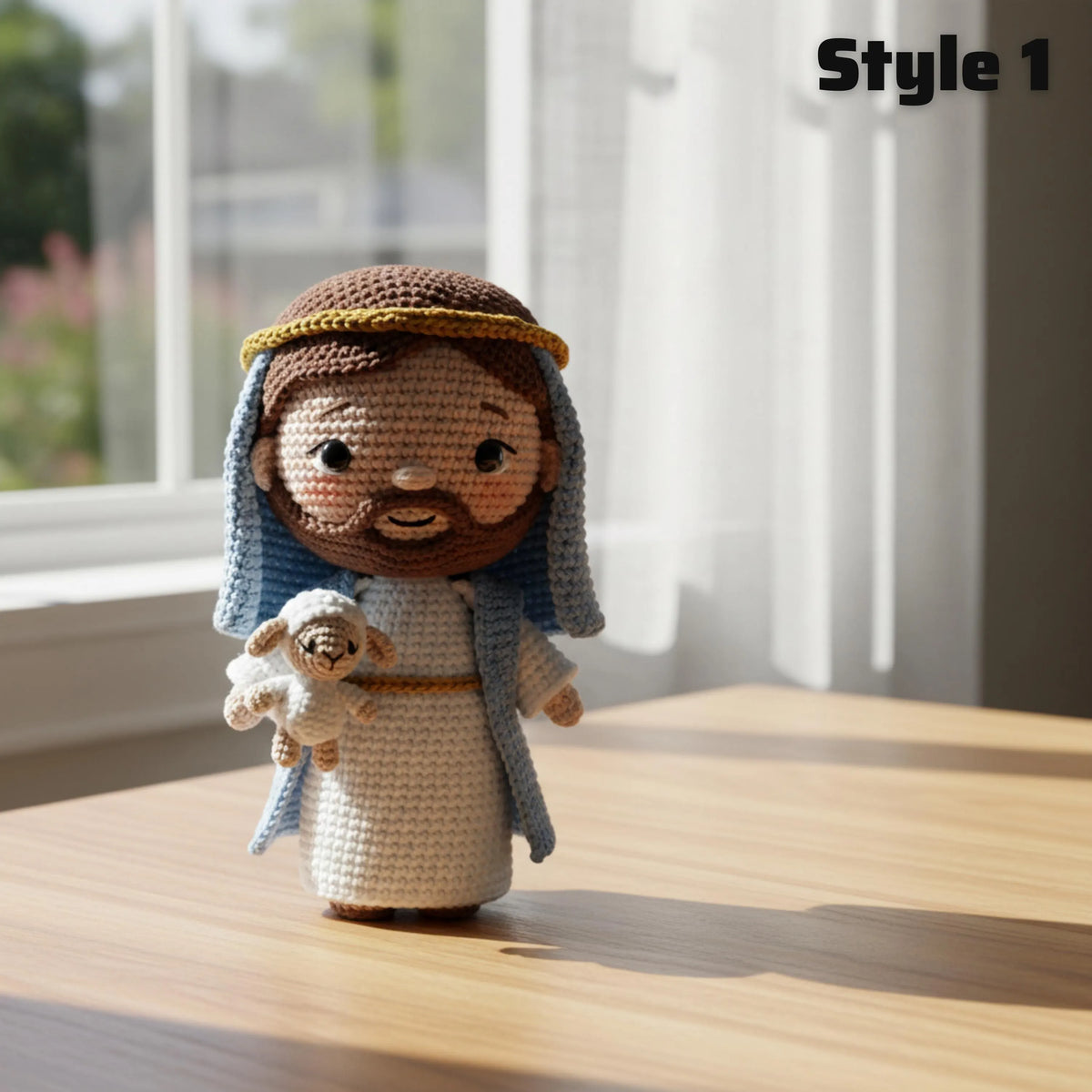 A crocheted doll of Jesus holding a lamb stands on a wooden table in a sunlit room. The doll has brown hair and beard, a white robe, a blue drape, and a gold headband.