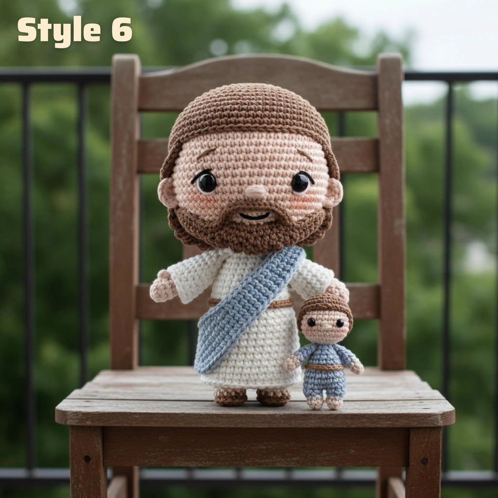Crocheted Jesus and child doll set styled on a wooden chair with a blurred outdoor garden background. The larger doll represents Jesus with brown hair and beard, white robe, and blue sash, while the smaller doll represents the child.