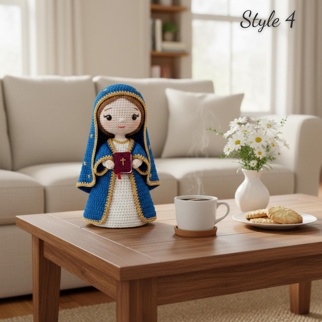 A crocheted doll dressed in a blue robe with gold trim and a white dress stands on a wooden coffee table in a living room setting. A white mug, a plate with cookies, and a vase of daisies are on the table, with a beige sofa and bookshelf in the background.