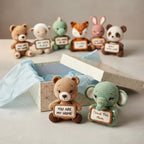 A set of crocheted plush animal figurines holding personalized sign plaques, displayed in front of and around an open gift box with tissue paper. The animals include a brown teddy bear, a white sheep, a green turtle, a fox, a pink rabbit, a panda bear, and a green elephant.