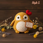 A crocheted penguin plush toy in mustard yellow and cream, holding a small wooden sign that reads 'Thank You For Making Me Who You Are,' styled on a dark wood surface with dried flowers and greenery. A red heart sits on its head, and the words 'Style 5' appear in the top right.