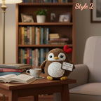 A crocheted owl plush toy sits on a wooden table in a cozy living room setting. The owl is brown with a white belly, yellow feet, and a small red heart on its head, holding a small sign that reads 'Thank You For Loving Me Best'.