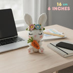 A crocheted white bunny plush holding an orange carrot with the word 'MOM' on it, placed on a light wood desk beside a laptop, notebook, and phone. The bunny wears a flower crown.
