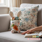 A crocheted white bunny plush holding an orange carrot sits on a light gray sofa in front of a floral throw pillow. A person's hand rests on an open book nearby.