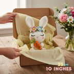 A crocheted white bunny plush holding an orange carrot, decorated with pastel flowers on its ears, displayed inside an open brown gift box lined with pale yellow tissue paper. Human hands are holding the box open, and a vase of flowers sits on the table beside it.
