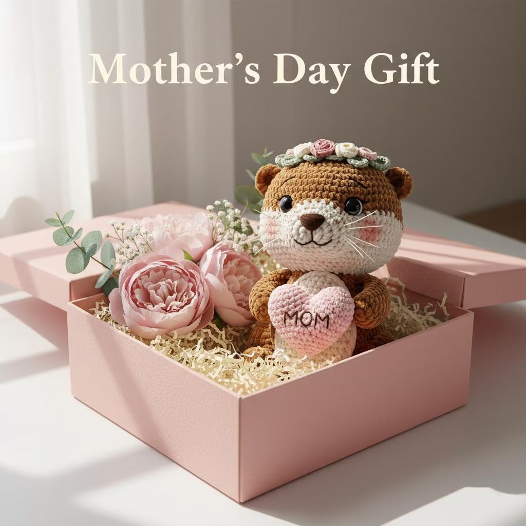 A crocheted brown animal plush toy with a floral crown and a pink heart that says “MOM,” displayed in a pink gift box with pink flowers and shredded paper. The text “Mother’s Day Gift” appears above on a soft neutral background.