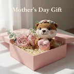 A crocheted brown animal plush toy with a floral crown and a pink heart that says “MOM,” displayed in a pink gift box with pink flowers and shredded paper. The text “Mother’s Day Gift” appears above on a soft neutral background.