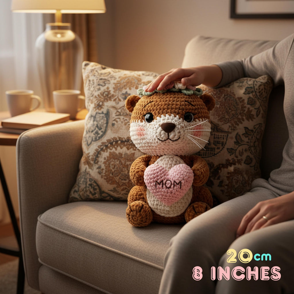 A crocheted otter plush toy with a pink heart that says 'MOM' sits on a beige armchair next to a person's hand and lap. The plush has brown and cream yarn, a small green leafy crown, and embroidered facial features.