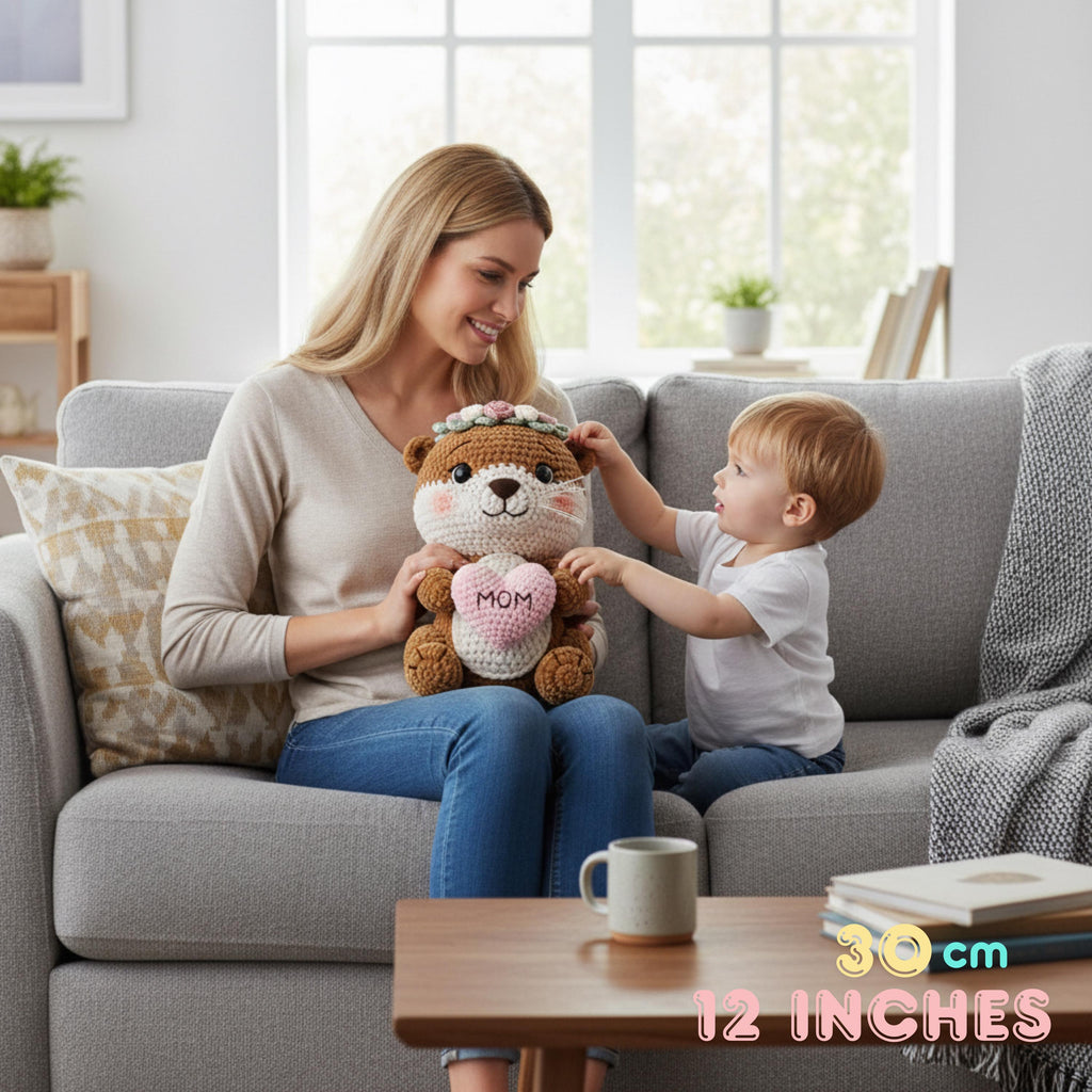 A woman and a young child sit on a gray sofa in a bright living room, holding a crocheted teddy bear plush with a pink heart that says “MOM.” The scene is styled as a cozy home interior with a coffee table, mug, pillows, and window in the background.