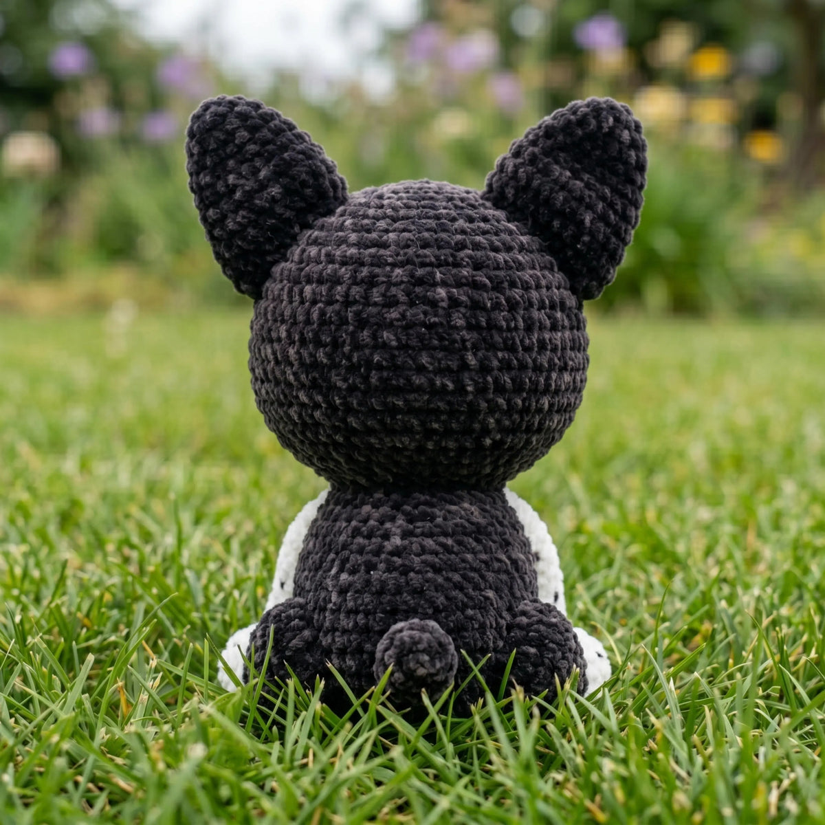 Back view of a handmade black and white crochet French Bulldog amigurumi doll sitting on green grass.