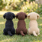 Three crocheted dog toys sitting on green grass outdoors, shown from the back. The toys are in three colors: black, brown, and cream.