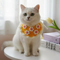 White cat wearing a colorful flower-patterned bib on a white surface with books and a tulip in the background.