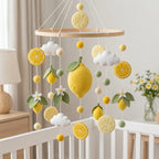 A baby nursery mobile featuring felt lemons, lemon slices, white clouds, and small white flowers hanging from a wooden ring over a white crib.