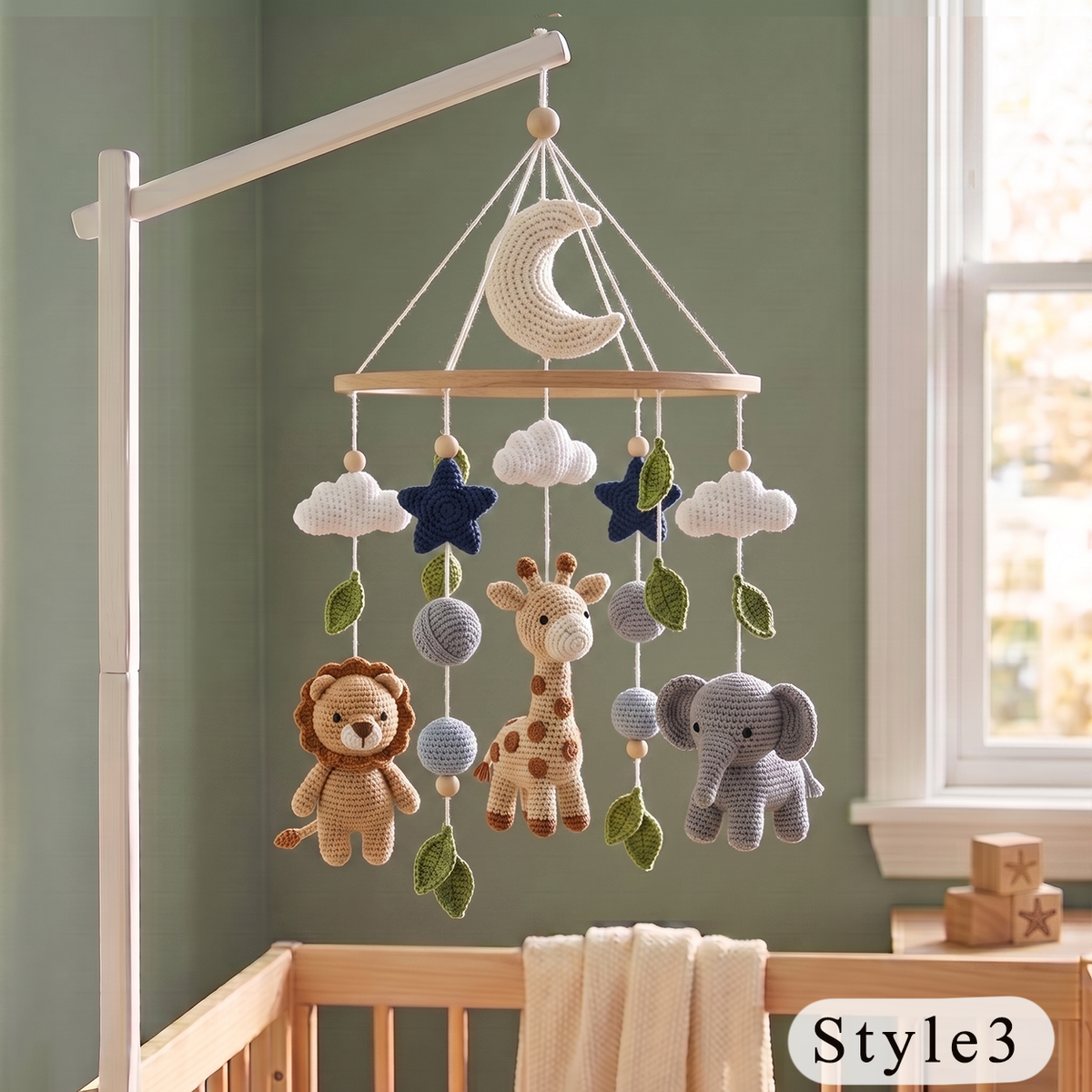 Crocheted baby mobile with a crescent moon, clouds, stars, lion, giraffe, and elephant hanging from a wooden bar on a white crib stand, set in a nursery with a green wall and window.