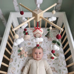 A baby in a cream onesie lies in a wooden crib, looking up at a hanging wooden baby mobile with crocheted Christmas-themed ornaments including a gingerbread house, tree, beads, and small plush figures.