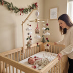 A baby in a wooden crib wearing a holiday-themed onesie, with a woman standing beside the crib. Above the crib is a wooden arch mobile decorated with white clouds, red pom-poms, green leaves, gingerbread houses, and a small Christmas tree.