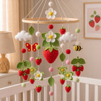 A strawberry-themed baby crib mobile hanging in a nursery. The wooden hoop mobile features felt strawberries, white flowers with yellow centers, green leaves, fluffy white clouds, a red-and-yellow butterfly, a bumblebee, and small pom-pom balls.