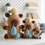 Three crocheted teddy bear toys in different sizes, featuring brown and tan bodies, cream-colored hats, and light blue teardrop-shaped noses, arranged on a white surface against a pegboard background.
