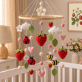 A baby crib mobile with felt strawberries, ladybugs, bees, clouds, and flowers hanging from a wooden ring in a softly lit nursery.